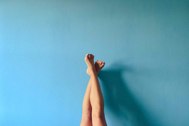 Woman’s legs crossed at the ankle, leaning against a blue wall.